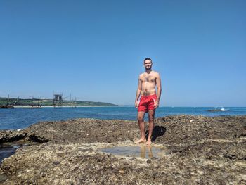 Full length of shirtless man standing on beach against clear sky