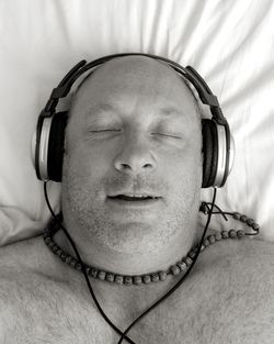 High angle view of shirtless man with eyes closed listening music on bed