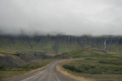 Country road leading towards grassy field against cloudy sky during foggy weather