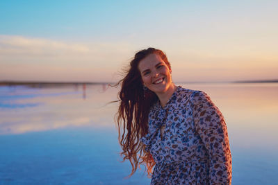 Portrait of smiling young woman standing against sea during sunset