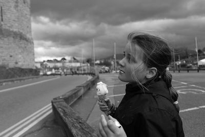 Side view of girl eating ice cream on road against cloudy sky