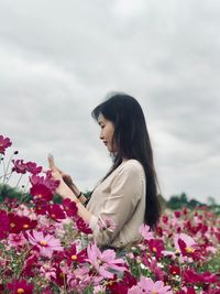 Side view of young woman photographing with mobile phone while standing on field against cloudy sky