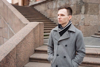 Beautiful model young guy in gray coat posing on city street during day.