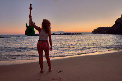 Rear view of woman with guitar standing on beach against sky during sunset