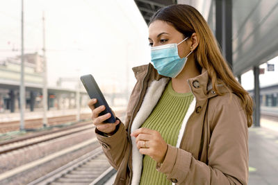Woman with surgical mask using her smart phone waiting her train at station outside