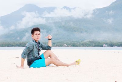 Portrait of young man sitting on beach against mountains