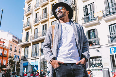 Adult hispanic male in trendy clothes with sunglasses and hat holding hands in pocket and looking away while standing on street