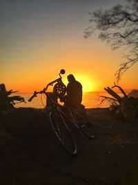 Rear view of man sitting by bicycle on shore against clear sky during sunset