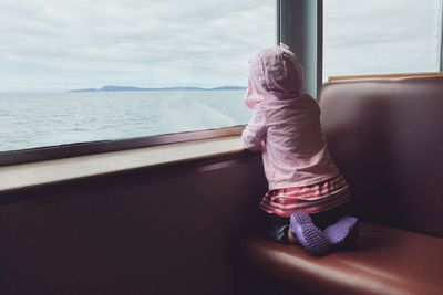 Rear view of girl looking through ferry window while wearing hooded shirt