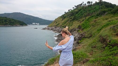 Rear view of young woman taking selfie while standing on mountain by sea against cloudy sky