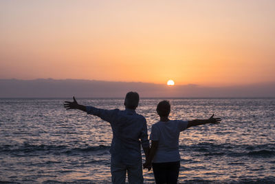 Rear view of couple with arms outstretched standing at beach against sky during sunset