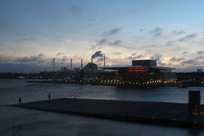 Illuminated city by river against sky at sunset