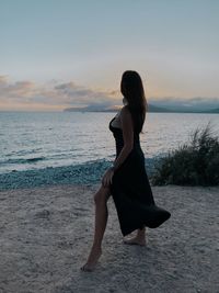 Rear view of woman standing at beach against sky during sunset