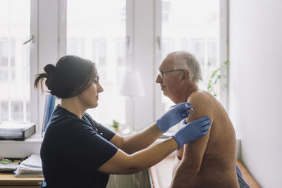 Female nurse giving vaccination to shirtless senior patient at hospital