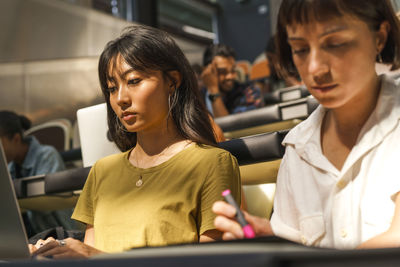 Low angle view of young female student using laptop while sitting by friend in classroom at university