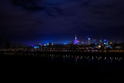 Illuminated buildings by river against sky at night