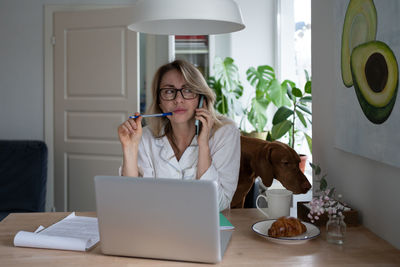 Woman with dog talking over phone by laptop on table