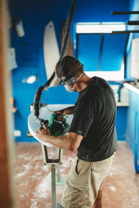 Worker in protective mask adjusting details surfboard in workshop