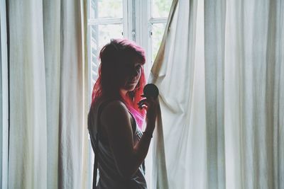 Woman with pink hair standing by curtain against window at home