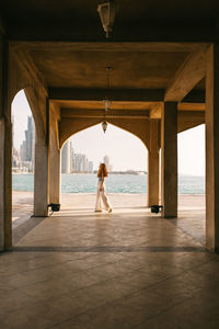 Woman walking under arch of traditional arabic building near sea while spending summer day in doha, qatar