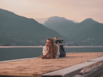 Full length of woman sitting by lake against mountains