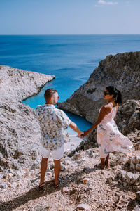 Rear view of couple standing on rock formation by sea against sky