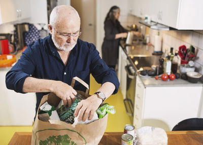 Senior man removing grocery from shopping bag while woman working in kitchen