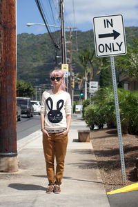 Full length of woman with hands in pockets standing by road sign in city