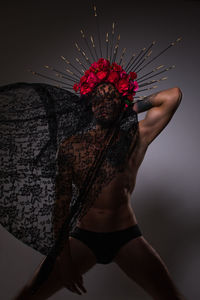 Shirtless man wearing headdress holding veil against gray background