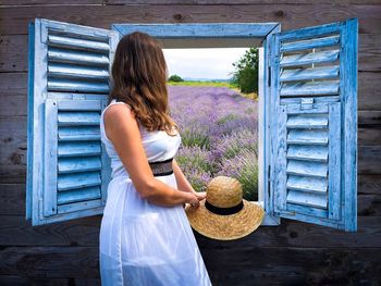 Woman in white dress looking out the open window at a field of lavender flowers