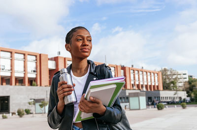 Pensive african american female student with thermos and bunch of textbooks looking away while standing on street near university building