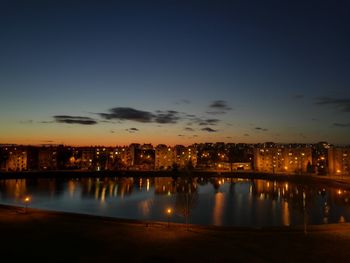 Illuminated city by river against sky at sunset