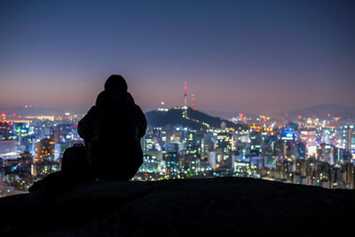 Rear view of man sitting against illuminated buildings in city at night