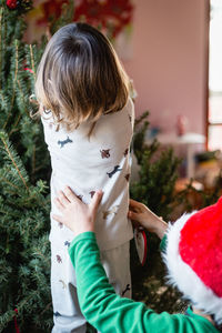 Back view of baby decorating christmas tree at home supporter by sister