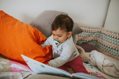 Cute baby boy reading book while sitting on bed at home