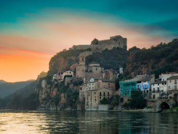 Buildings by lake against sky during sunset