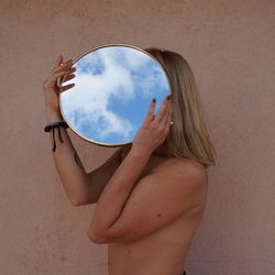 Shirtless woman holding mirror standing against wall
