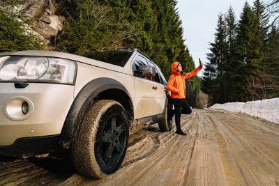 Man in orange jacket taking selfies and standing by off-road car on muddy mountain rroad in winter