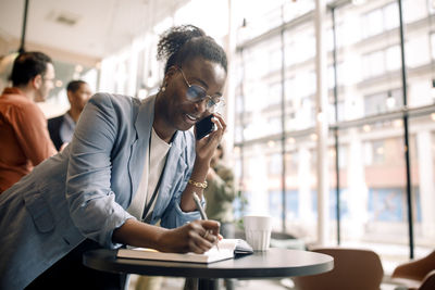 Smiling businesswoman preparing notes while talking through smart phone at convention center