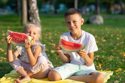 Brother and sister holding watermelon while sitting in park