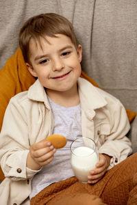 Little adorable boy sitting on the couch at home, drinking milk with cookie. fresh milk in glass