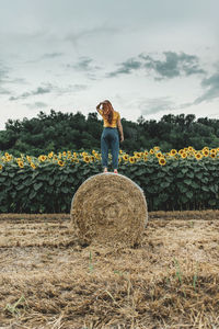 Full length of young woman standing on hay bale in front of a sunflowers field
