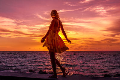 Full length of beautiful woman walking on retaining wall by seascape against sky during sunset