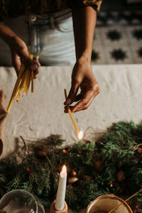 From above of crop anonymous female with bunch of candles lighting up in hands standing near table with various xmas decorations while preparing for celebration at home