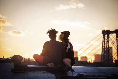 Cheerful couple sitting on retaining wall during sunset