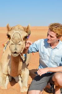 Young man petting camel on desert against clear sky during sunny day