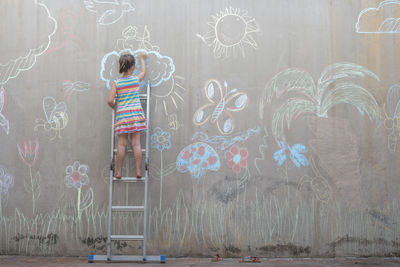 Girl standing on ladder drawing colourful pictures with chalk on a concrete wall