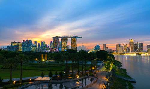 Illuminated buildings by river against sky at sunset