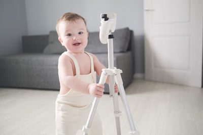 European child of 1 year in white jumpsuit with tripod for photo of house in bright real interior of 