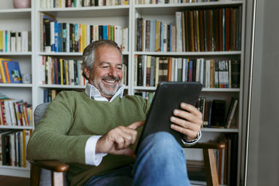 Smiling man using digital tablet while sitting against bookshelf at home